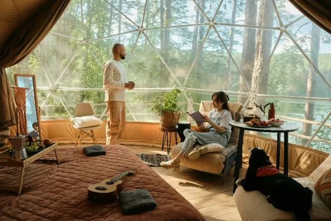 Couple relaxing in a cozy glamping dome with forest views, snacks, and their dog.